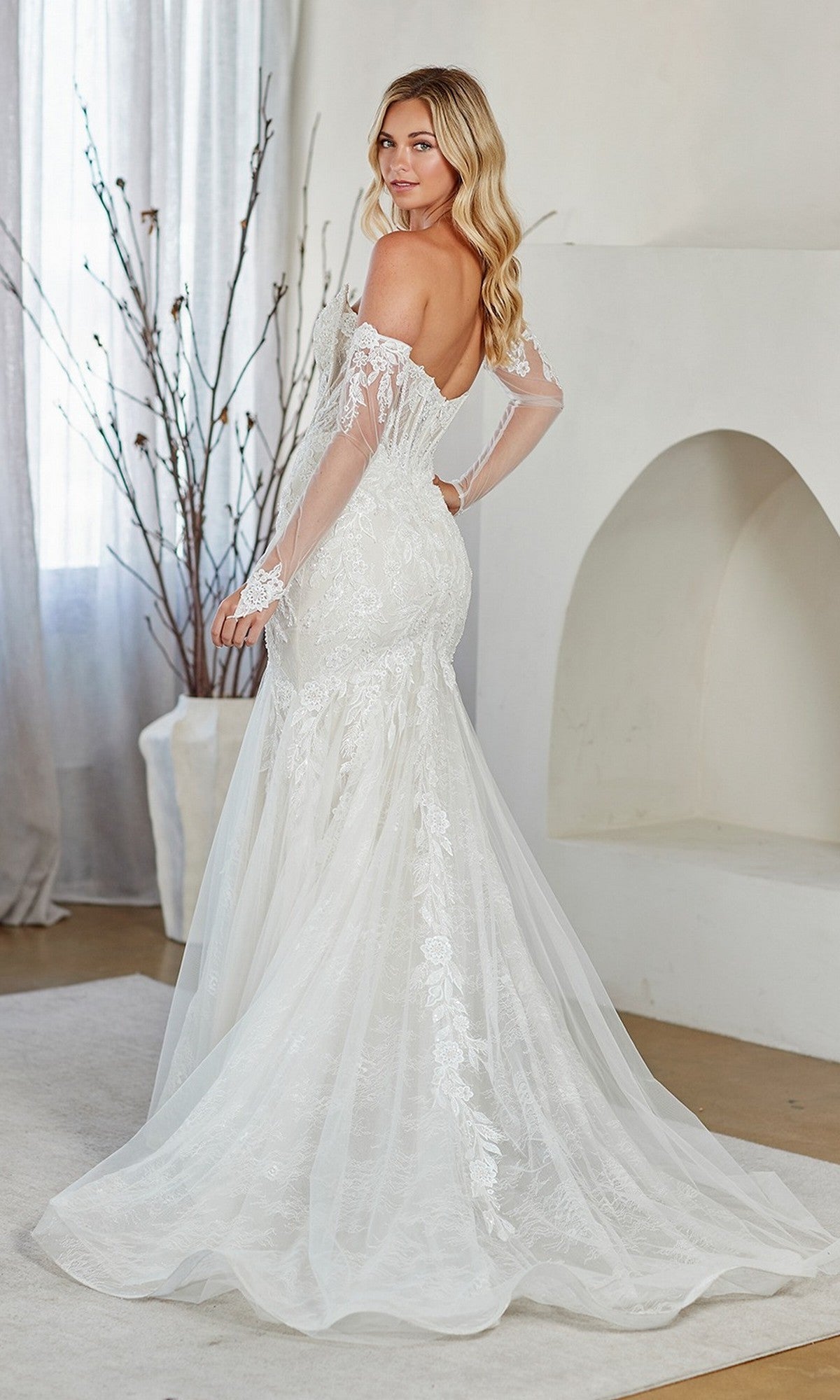 A woman in the Amelia Couture 2155 long prom dress, featuring white lace embroidery, a strapless corset bodice, and sheer off-the-shoulder sleeves, stands indoors and looks over her shoulder.